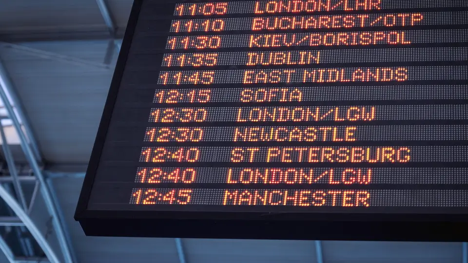 An airport departures board with orange dot-matrix text listing flight times and destinations, including Edinburgh, Dublin, Sofia, Newcastle, Manchester and several London airports.