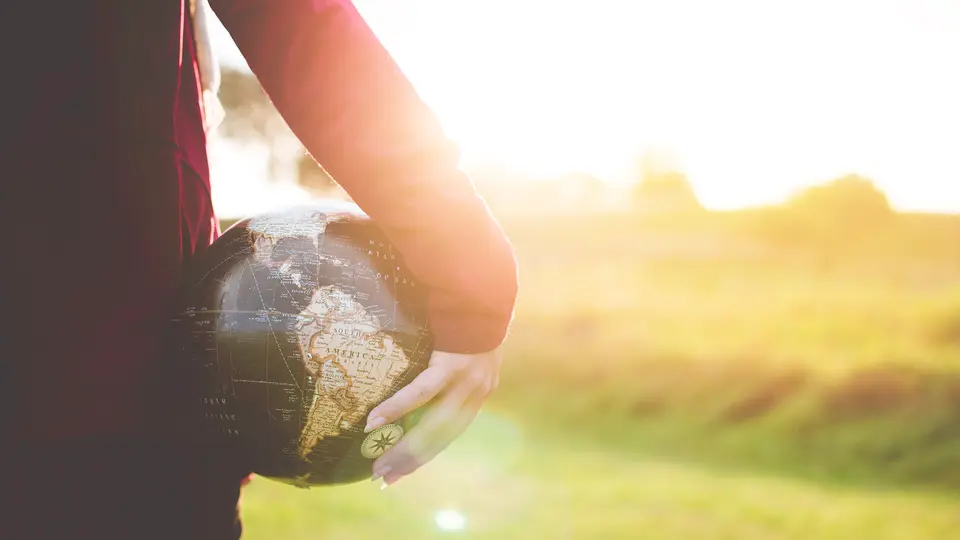 A person in a long-sleeved burgundy top holding a dark vintage globe against their hip, with South America facing out, standing in a sunlit grassy field with trees blurred in the background.