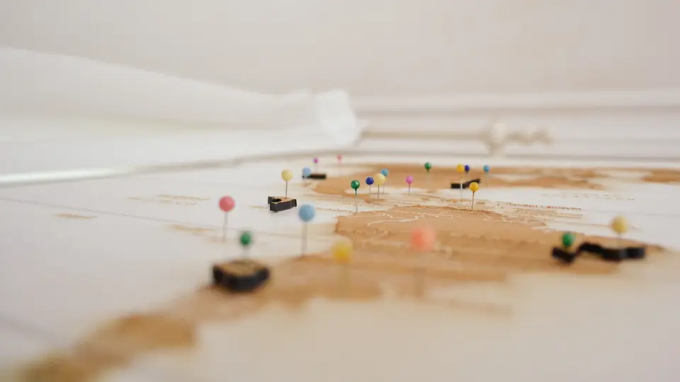 A wooden world map laid flat on a white surface, with coloured push pins marking places across several continents and the far edge softly out of focus.