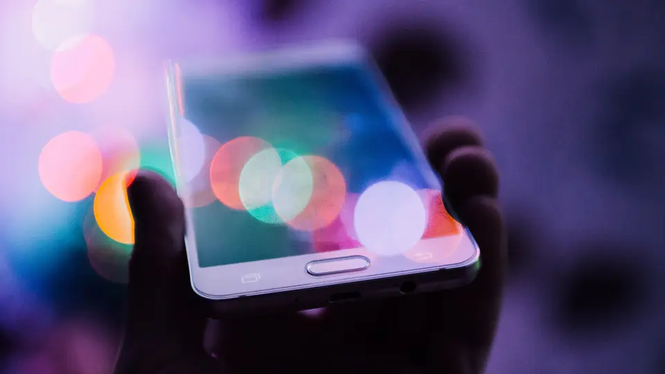 A hand holding a white smartphone at an angle, its screen glowing with soft out-of-focus circles of pink, orange and blue light against a dark purple background.