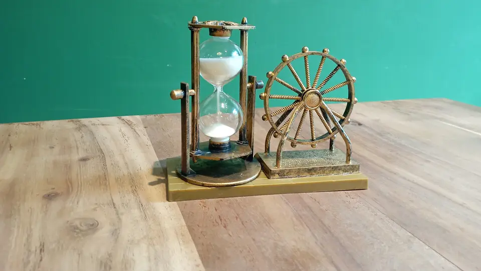 A small brass hourglass with white sand running into the lower bulb, standing next to a miniature brass Ferris wheel on a wooden table in front of a green wall.
