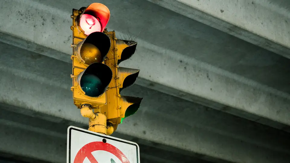 A yellow traffic light mounted under a concrete overpass, showing a red light at the top with the amber and green lights unlit, above the top of a white road sign with a red prohibition circle.