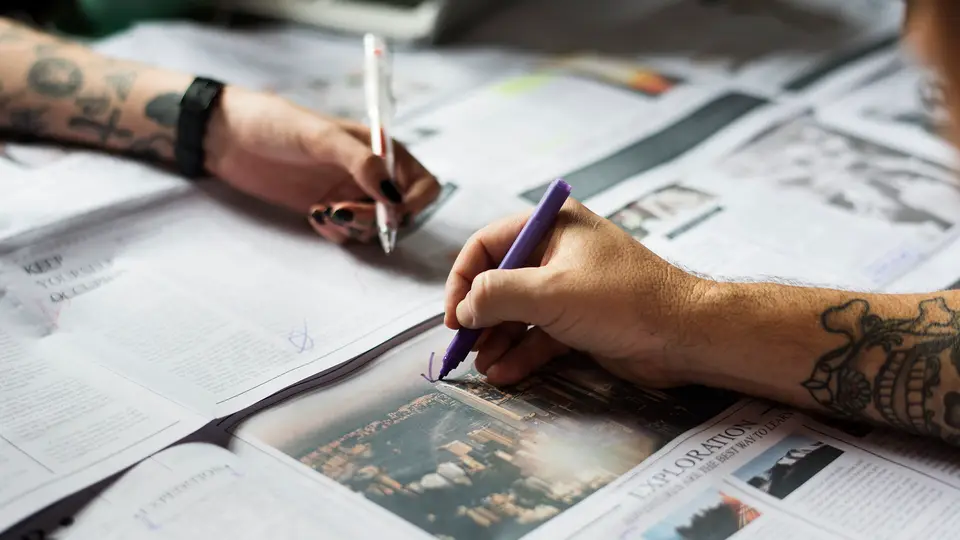 Two people with tattooed forearms marking up spread-out newspaper pages with pens.