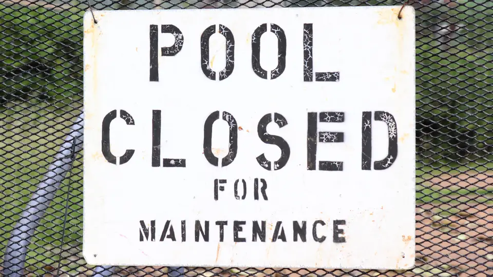 A weathered white metal sign with black stencilled letters reading Pool closed for maintenance, hung on a chain-link fence with blurred greenery behind it.