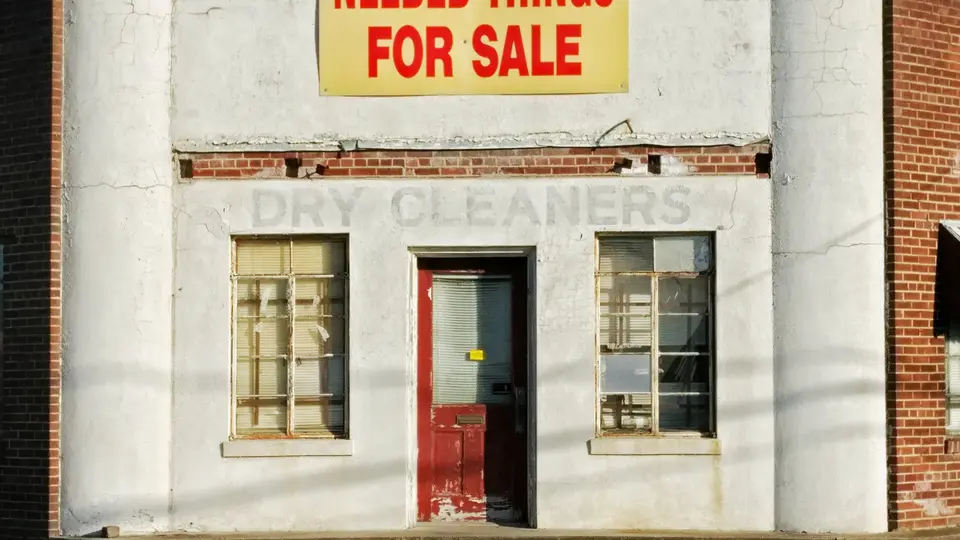 A white-painted brick shopfront with a red door, two multi-pane windows, a faded painted Dry Cleaners name above them and a yellow For Sale banner at the top.