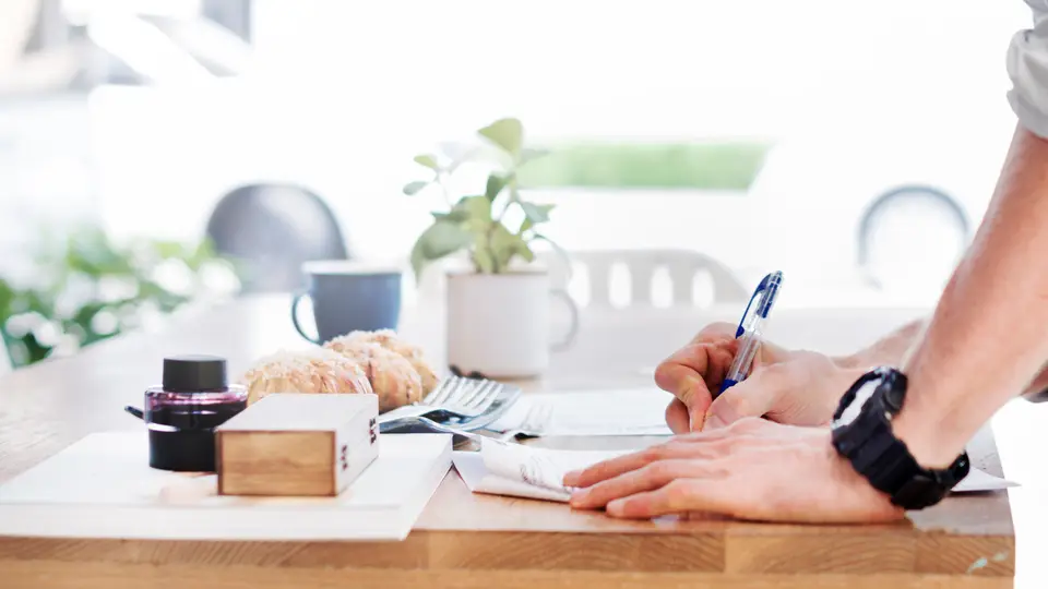 A man in a grey shirt leaning over a wooden table, writing on sheets of paper with a blue pen, beside a small potted plant, a coffee cup and a tray of pastries.