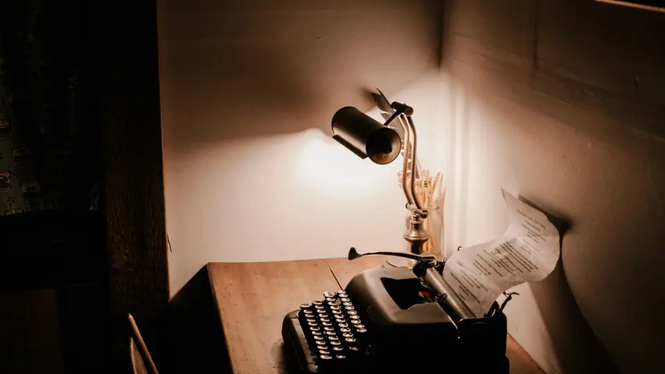 A dark grey vintage typewriter on a wooden desk beneath a brass desk lamp, with a typed sheet of paper curling out of the roller and warm light falling on a beige wall.