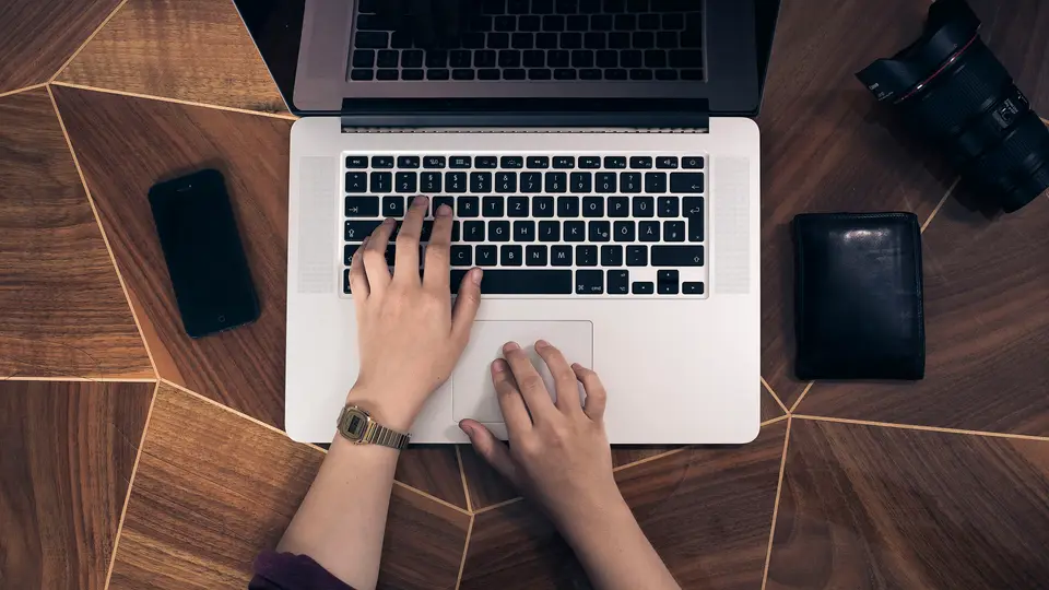 Top-down view of two hands typing on a silver laptop keyboard on a geometric wooden table, with a smartphone, a black wallet and a camera lens beside the laptop.