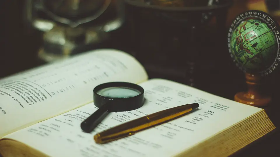 A black magnifying glass and a gold pen resting on the pages of an open book, with a small green desk globe in the background.