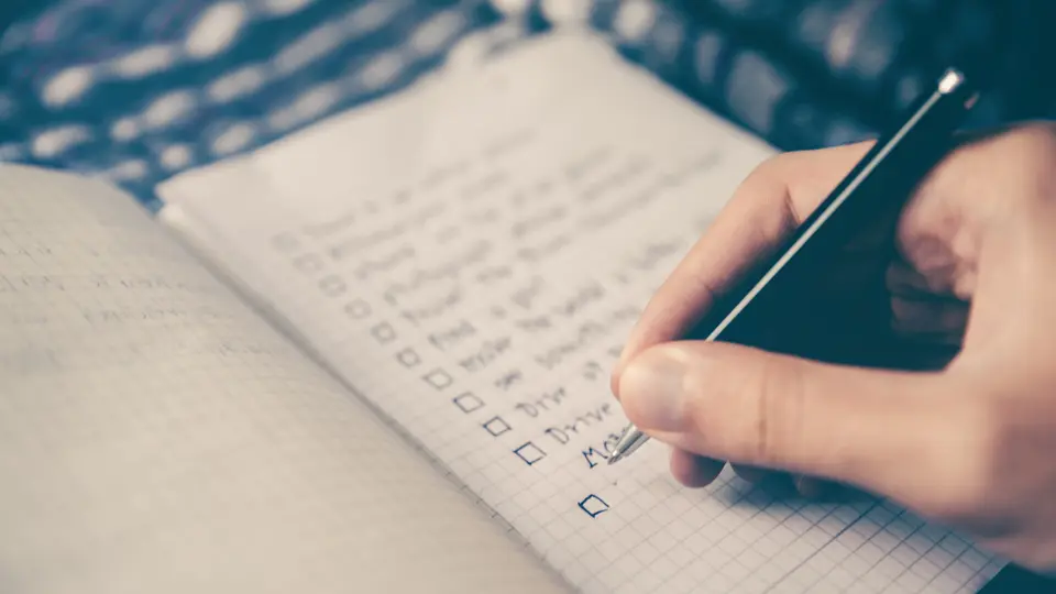 A hand holding a black pen over a grid-paper notebook, writing a handwritten to-do list with an empty tick box beside each task.
