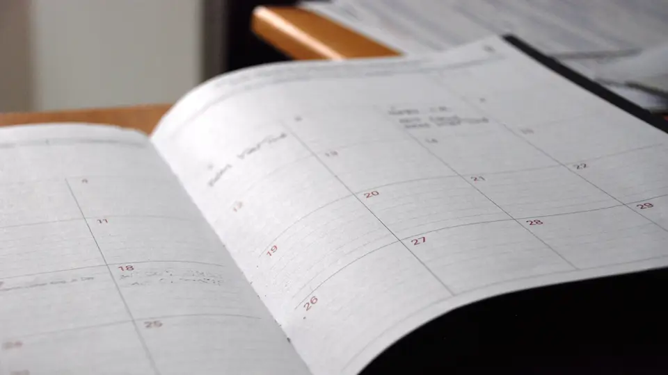 An open paper diary on a wooden desk showing a monthly calendar grid with red date numbers and a few handwritten notes in grey pencil.