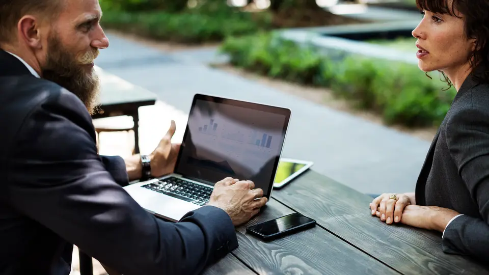 A bearded man in a suit points at his laptop screen while talking with a woman at an outdoor table.