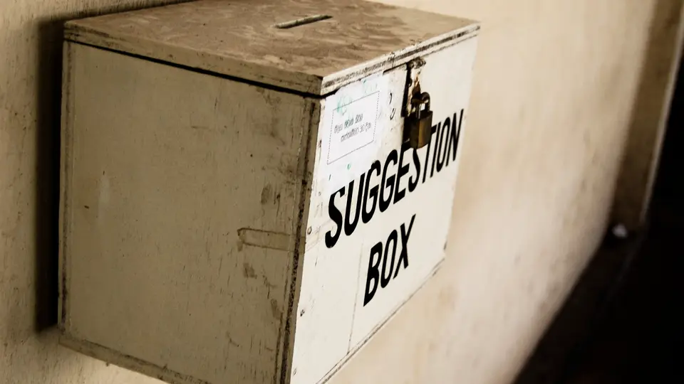 A worn white wooden suggestion box with a padlock and a slot in the lid, mounted on a plain cream wall