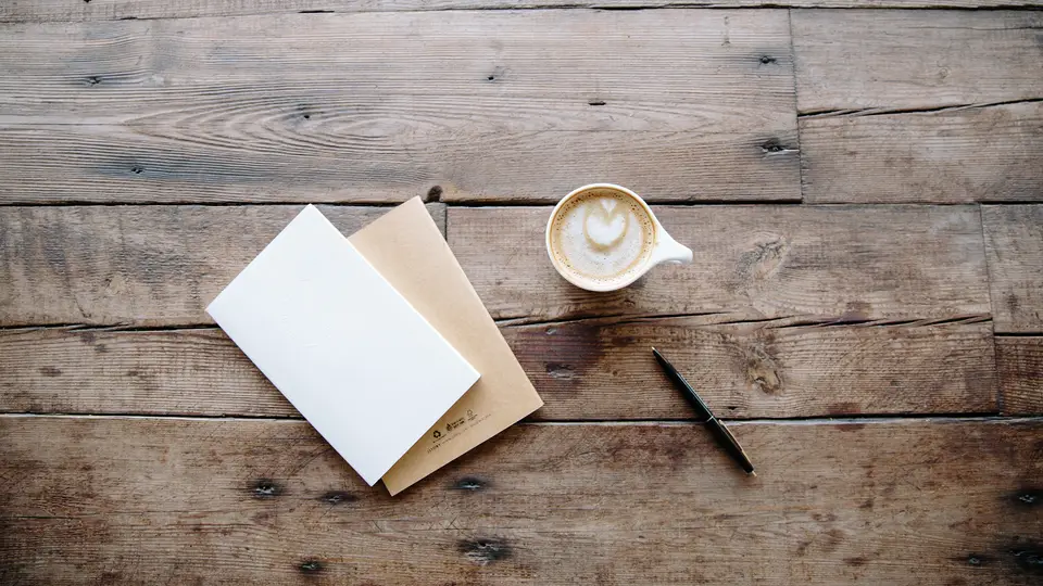 Two closed notebooks, a pen and a cup of latte on a rustic wooden table, seen from above, ready for planning.