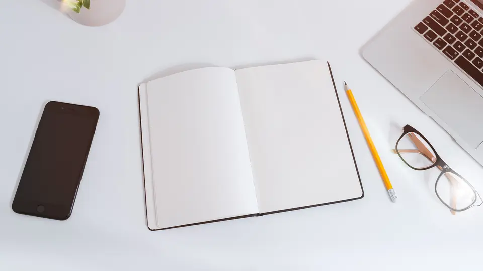 A white desk seen from above with an open blank notebook, a yellow pencil, a pair of glasses, a smartphone, a small potted plant and the corner of a laptop keyboard.