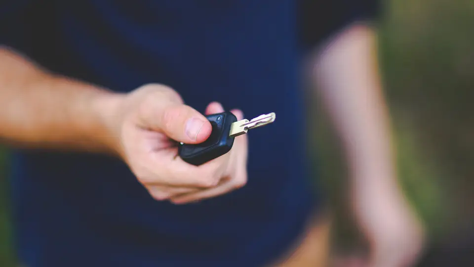 A hand holding out a black key fob and a metal key towards the camera, as if handing it over, against a blurred dark blue background.