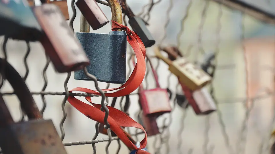 Several old, weathered padlocks of different sizes locked onto a wire mesh fence, one tied with a faded red ribbon.