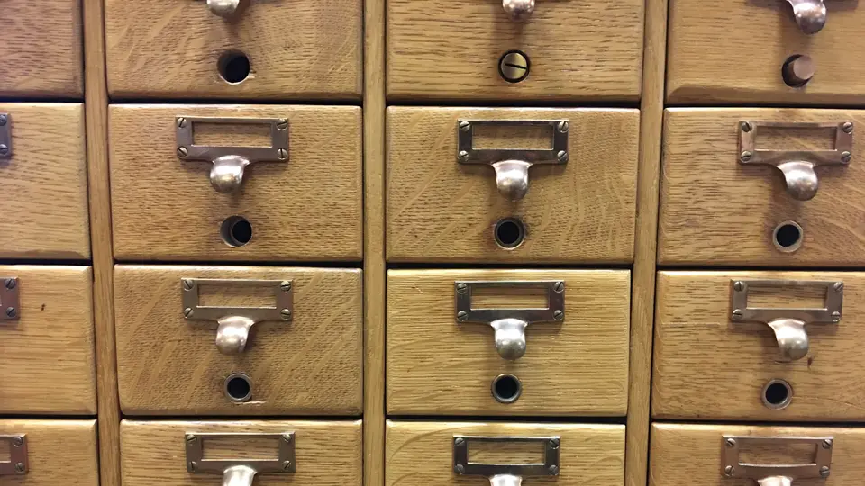 Rows of honey-coloured oak card catalogue drawers, each with a metal pull handle and an empty label holder, filling the frame
