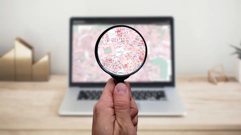 A hand holding a round magnifying glass up to a laptop screen, enlarging part of a detailed property map, with the laptop on a pale wooden desk.
