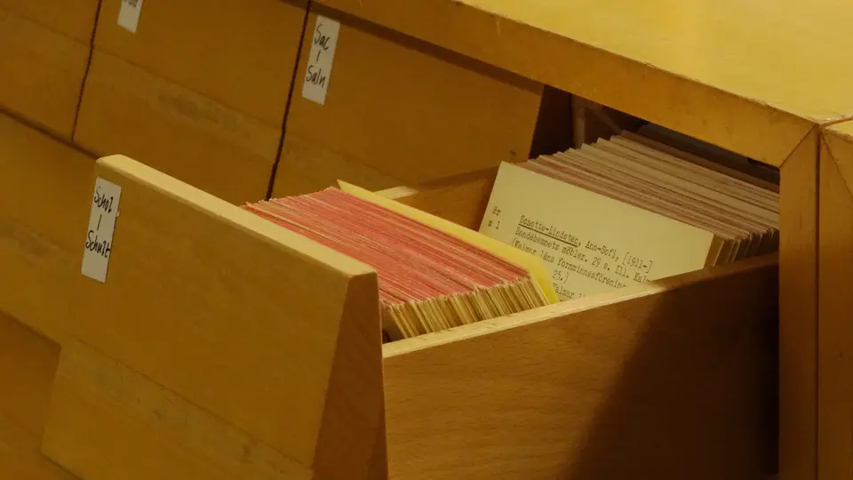 An open pale wooden card catalogue drawer packed with pink-edged and cream index cards, next to drawers marked with small handwritten labels.